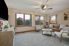 A spacious living room with a ceiling fan, large windows, a wooden desk, and a leather sofa.