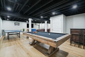 A pool table with a billiard cue rack and balls, a sofa, and a door in a room with wooden flooring and exposed ceilings.