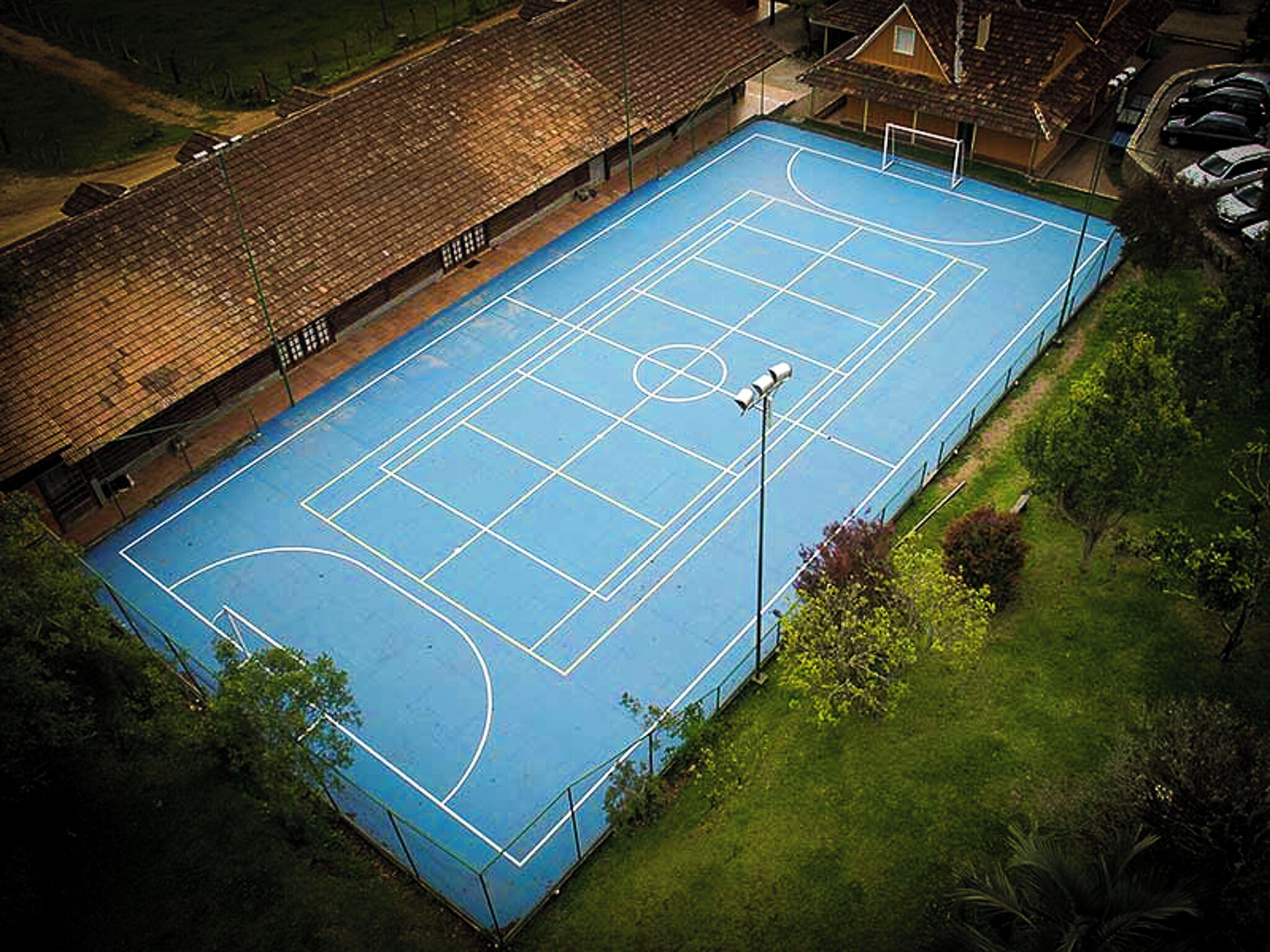 Una cancha de baloncesto al aire libre con superficie azul, rodeada por una valla y contigua a un edificio de techo inclinado.