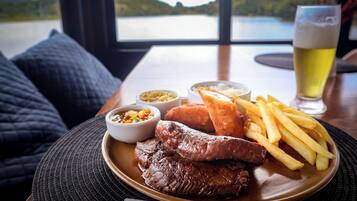 Un piatto con bistecca, patatine fritte e pane, accompagnato da una birra e salse su un tavolo vicino a una finestra con vista su un lago.
