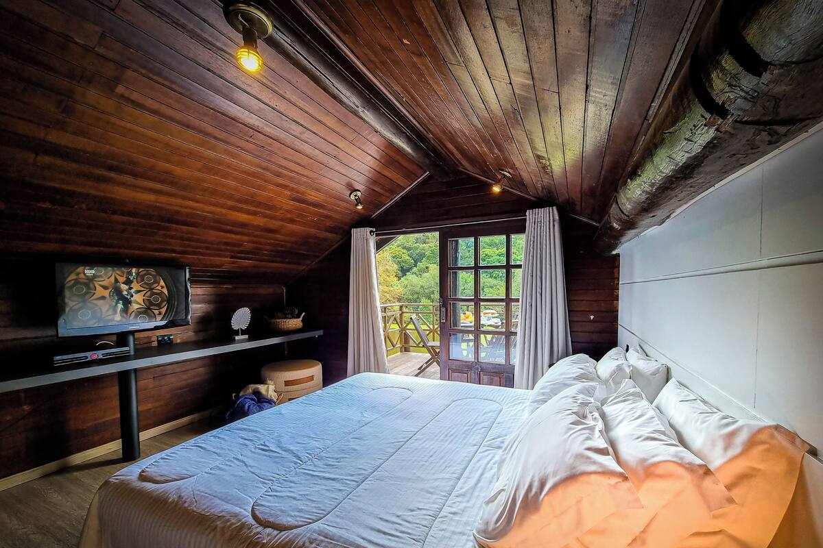 Una camera da letto con soffitto in legno, un letto con biancheria bianca, una televisione su uno scaffale e una finestra con tende.