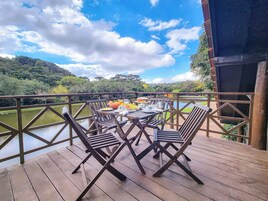 Una terraza de madera con una mesa dispuesta para el desayuno, con vista a un lago y colinas boscosas.