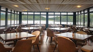 Een restaurant met grote ramen en uitzicht op een meer. Tafels en stoelen staan klaar voor het dineren.