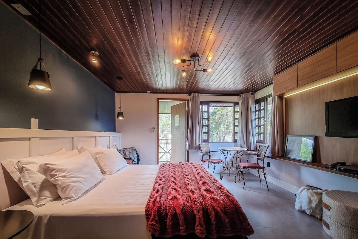 Una camera da letto con soffitto in legno, un letto con lenzuola bianche, una coperta rossa, una TV e un angolo ristoro con sedie e tavolo.