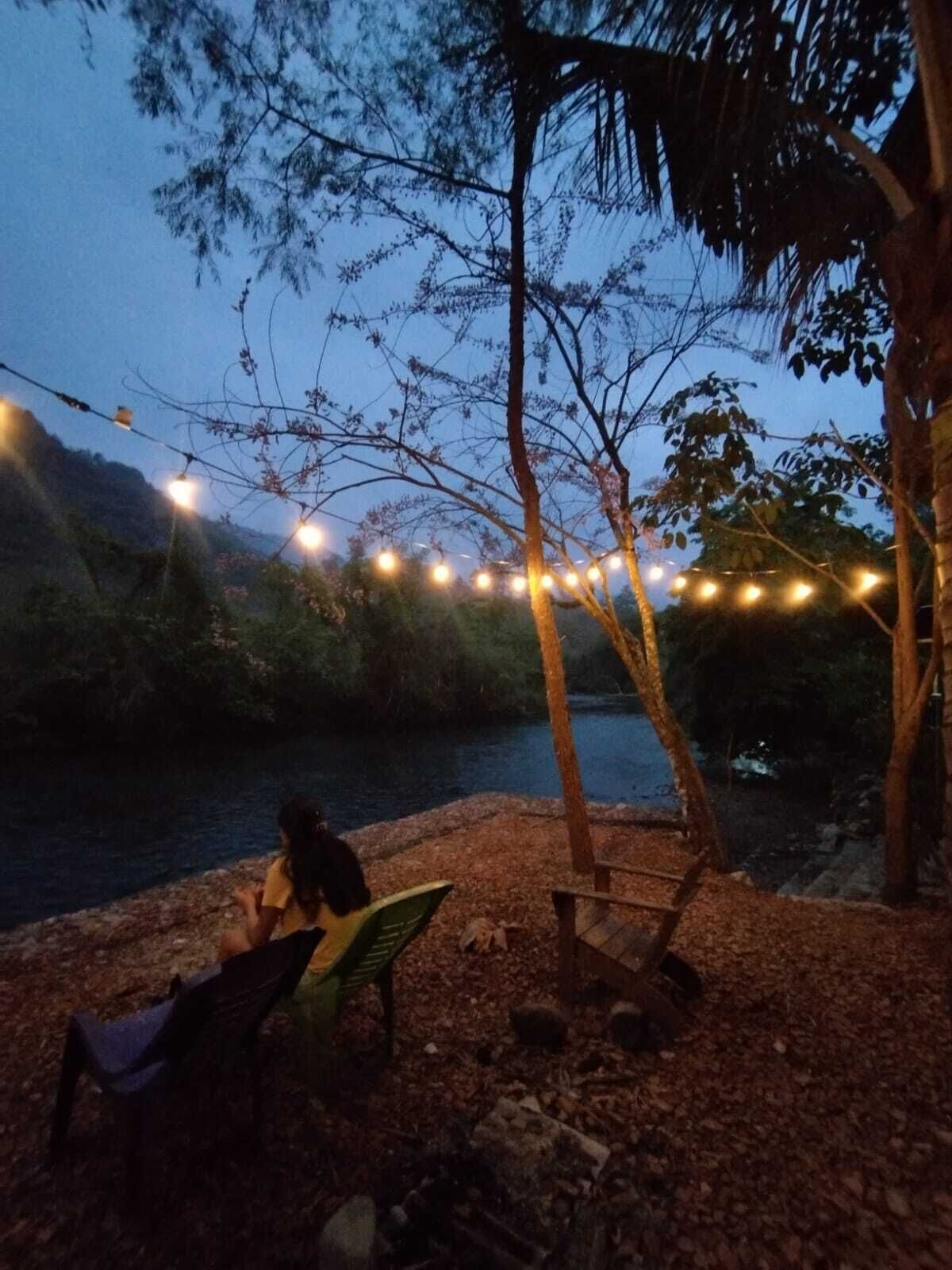 A riverside setting with string lights, a bench, and a person reading a book.