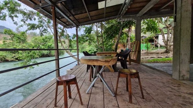 A wooden deck with a view of a river, surrounded by greenery, and featuring a covered seating area with tables and chairs. - Yaxlik River lodge (San Agustin Lanquin)