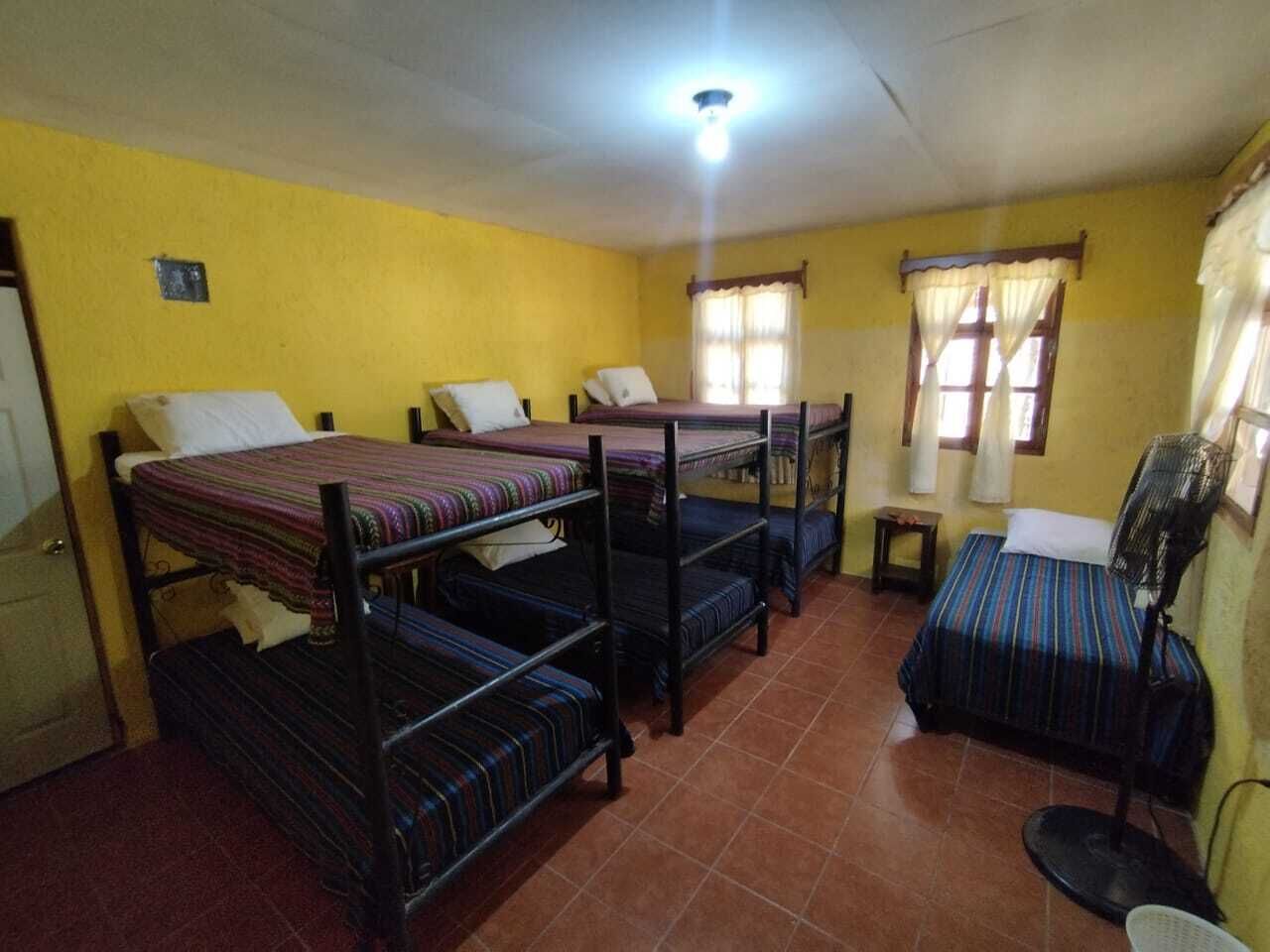 A dormitory room with four bunk beds, a small table, a chair, and a standing fan.