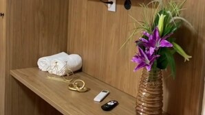 Ett rum med en väggpanel av trä, en väggmonterad tv, ett litet vitt kylskåp, ett skrivbord i trä, en vas med blommor och en fjärrkontroll.