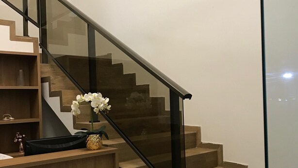 En modern trappa med glasräcken och trästeg, som leder till en reception med ett träbord och en vas med vita blommor.