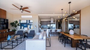 A spacious living area with a sofa, coffee table, and dining table. A ceiling fan and pendant lights are visible. A kitchen area is partially visible in the background. - Luxury condo with pool, jacuzzi and private terrace (Cabo San Lucas)