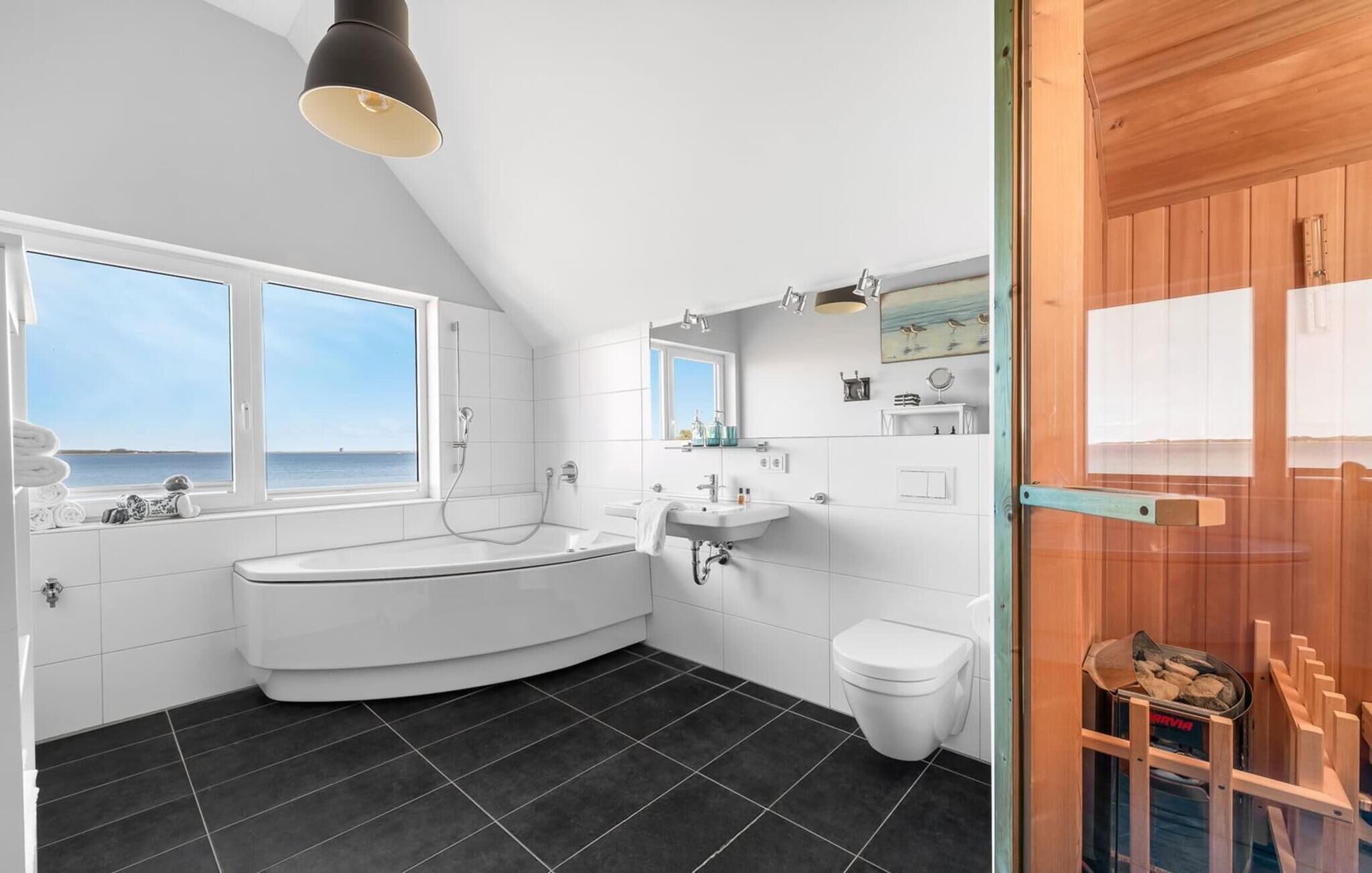 A modern bathroom with a bathtub, a sink, and a toilet. There are two windows providing a view of the sea.