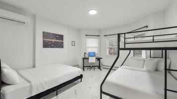 A dorm-style room with bunk beds, a desk with a computer, a window, and a wall-mounted air conditioner.