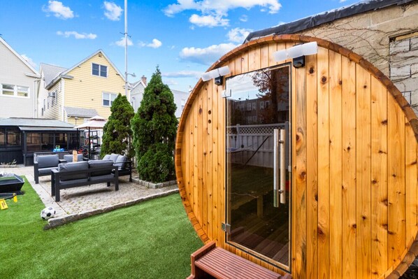 A wooden sauna with a glass door, a patio with outdoor furniture, and a well-maintained lawn.