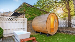 Eine Sauna mit Holzfassade, eine weiße Außendusche und eine hölzerne Bank.