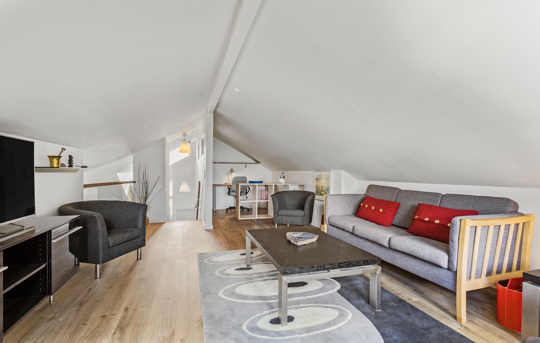 Eine moderne Loft-Wohnung mit hohen Decken, einem grauen Sofa mit roten Kissen, einem Holzstuhl, einem schwarzen Schreibtisch und einem grauen Teppich mit Muster.