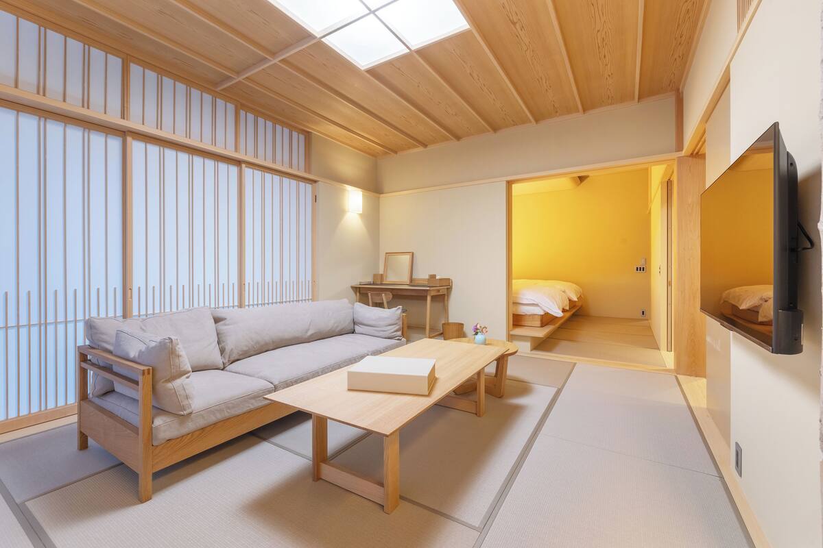 A traditional Japanese-style room with tatami flooring, a low wooden table, a sofa, and a television mounted on the wall.