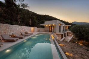 Un espace moderne aménagé au bord de la piscine, avec des chaises longues, un mur en pierre et un bâtiment doté d’un balcon.