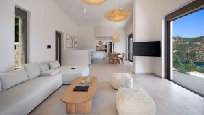A modern living room with a sofa, coffee table, and ottoman, a dining area with chairs, and a kitchen in the background. - Villa Eptanisa in Sivota (Lefkada)