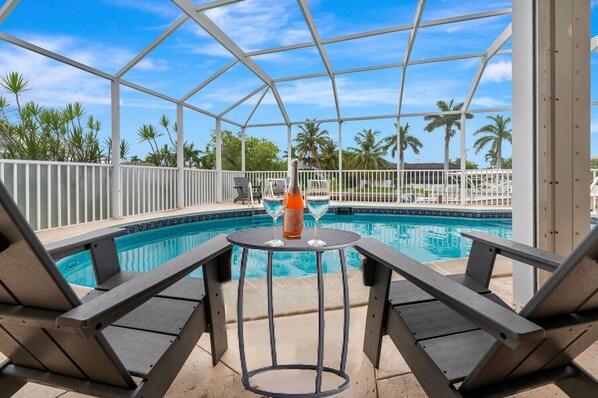 A pool area with a covered seating area, a table with a bottle and glass, and palm trees in the background.