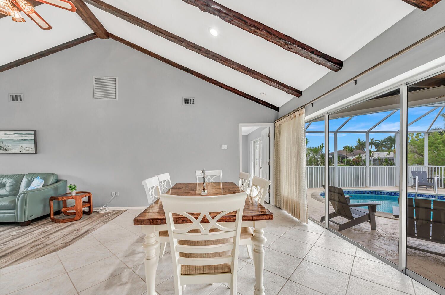 A spacious living room with a wooden dining table, white chairs, and a green sofa. A glass door leads to an outdoor pool area.