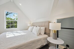 A bedroom with a large bed, a bedside table with a lamp, a window with a view of trees, and a sloped ceiling. - Modern New 1-bedroom Guest House for 3 or Combined with Main House for 9 people  (Houston)