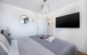 Et moderne hotelværelse med en stor seng, et fladskærms-tv, et rundt spejl og et lille bord med en lampe og blomster.