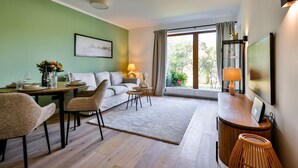 A modern living room with a sofa, dining table, and chairs. There is a large window with curtains, a lamp, and a television on a stand. - Lille, Wenningstedt-braderup auf Sylt, Deutschland (Wenningstedt-Braderup (Sylt))