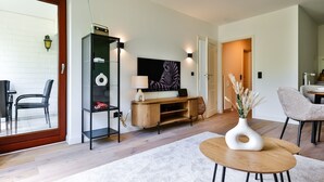 A modern living room with a glass door leading to a balcony, a wooden entertainment unit, a large flat-screen TV, and a dining area with a table and chairs. - Lille, Wenningstedt-braderup auf Sylt, Deutschland (Wenningstedt-Braderup (Sylt))