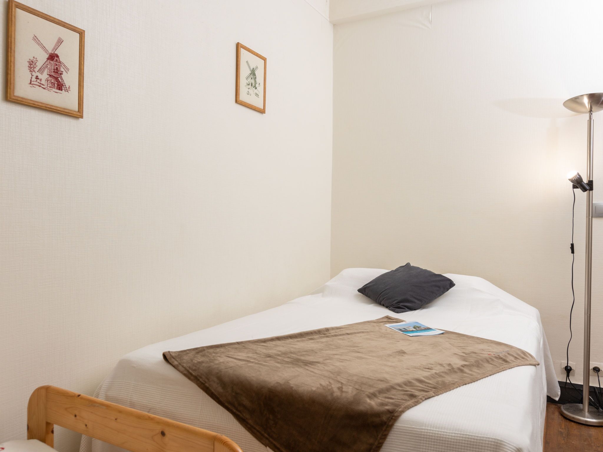 A neatly made bed with a brown blanket, a black pillow, and a small picture on the bed. There is a wooden chair and a floor lamp beside the bed. Two framed pictures are hung on the wall above the bed.