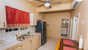 A compact kitchen with wooden cabinets, a black refrigerator, a ceiling fan, and a dining area with a brown table and yellow chairs. - Martin's Way@Potters Village 2 BR Apartment (Potters Village)