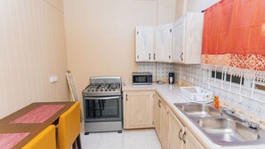 A compact kitchen with a gas stove, microwave, and double sink. There is a dining table with yellow chairs and a red placemat. - Martin's Way@Potters Village 2 BR Apartment (Potters Village)