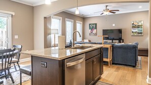 A modern kitchen with a central island, a dining area with a table and chairs, a living room with a sofa and TV, and a ceiling fan. - Mulligan Manor- 0.2 mile walk to Vitos Ristorante and Pizzeria and many others! (Southern Pines)
