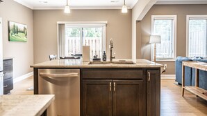 A kitchen with a central island, a dining area with a blue sofa, and a view of a deck through large windows. - Mulligan Manor- 0.2 mile walk to Vitos Ristorante and Pizzeria and many others! (Southern Pines)