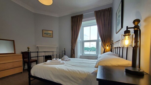 A neatly made bed with white linens, a wooden dresser, a bedside lamp, and a window with curtains. - Little Bay View in Oban (Oban)