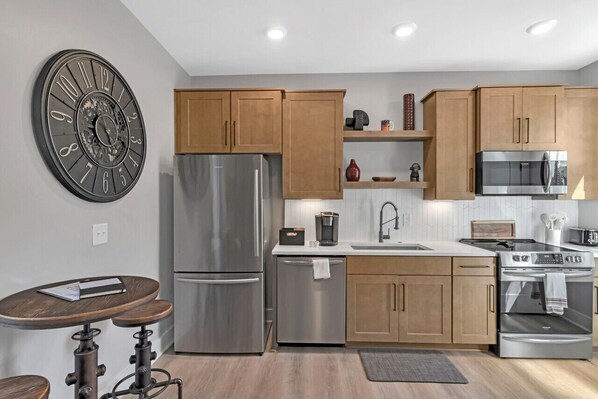A modern kitchen with wooden cabinets, stainless steel appliances, a round clock, and a small dining table. - Roof Deck|Pet Friendly|E.Nash (Nashville)