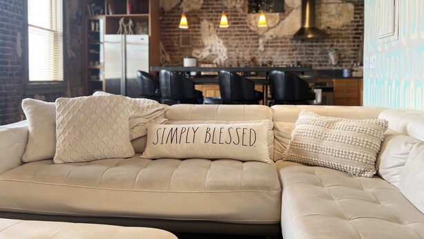 Un espacio de sala acogedor con un sofá color crema, almohadones estampados y una cocina al fondo.