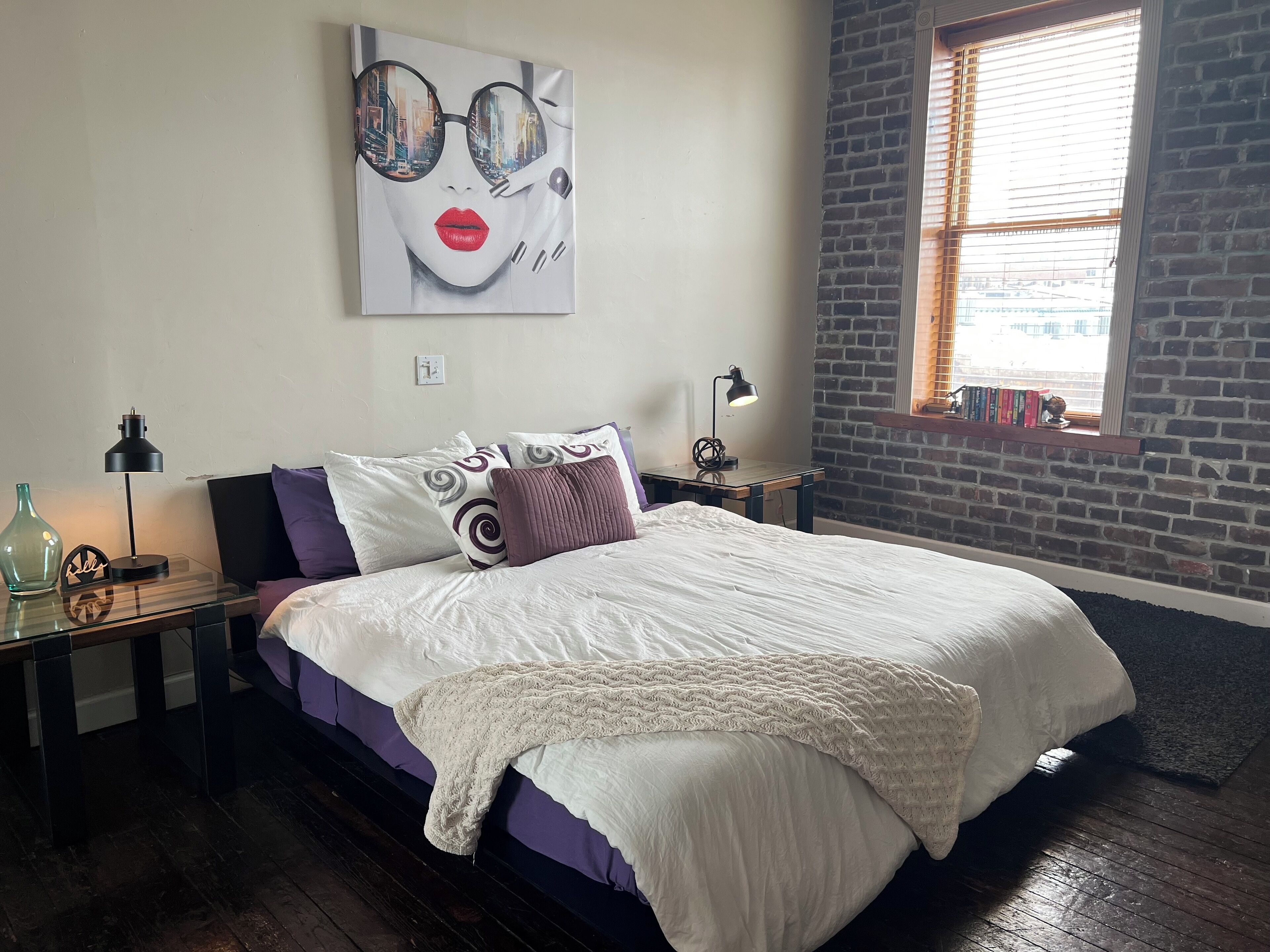 Un dormitorio con una cama, dos mesitas de noche, un reloj de pared, una ventana con persianas y una pared de ladrillo.