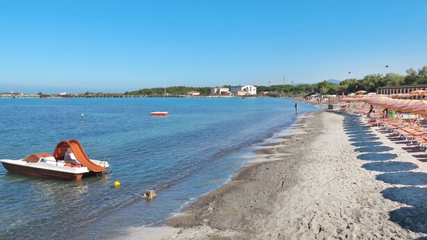 Een strand met een rij ligbedden en parasols, een boot, en een helderblauwe lucht.