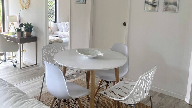 A modern living room with a round white table, white chairs, a woven ceiling light, and a door with a beach-themed sign. - Welcome to our serenity in the Gold Coast which is "Bee Cottage".
 (Robina)