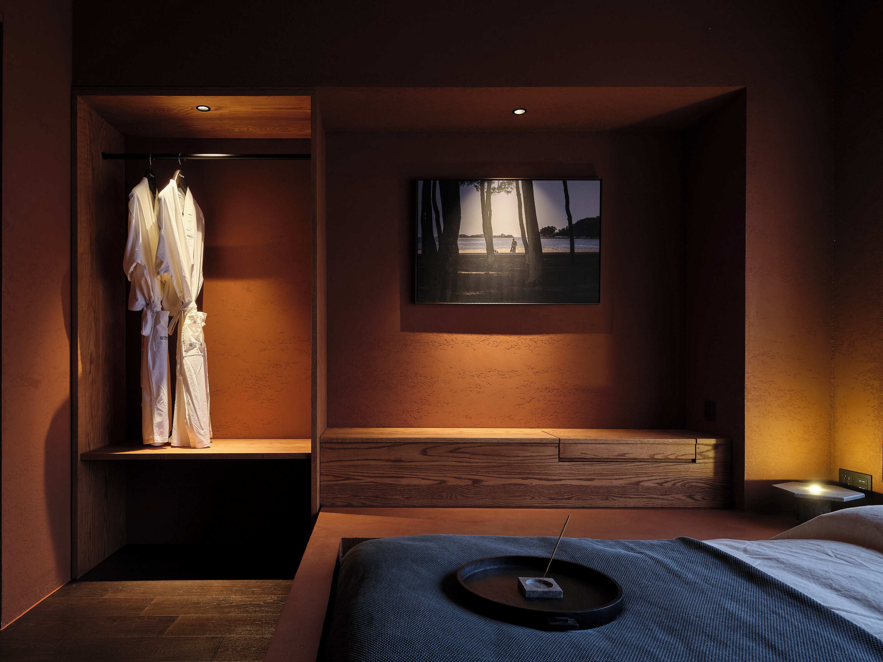 Un dormitorio moderno con un armario de madera, una cama con cabecero oscuro, una mesita de noche pequeña y un cuadro colgado en la pared que representa un atardecer sobre árboles.