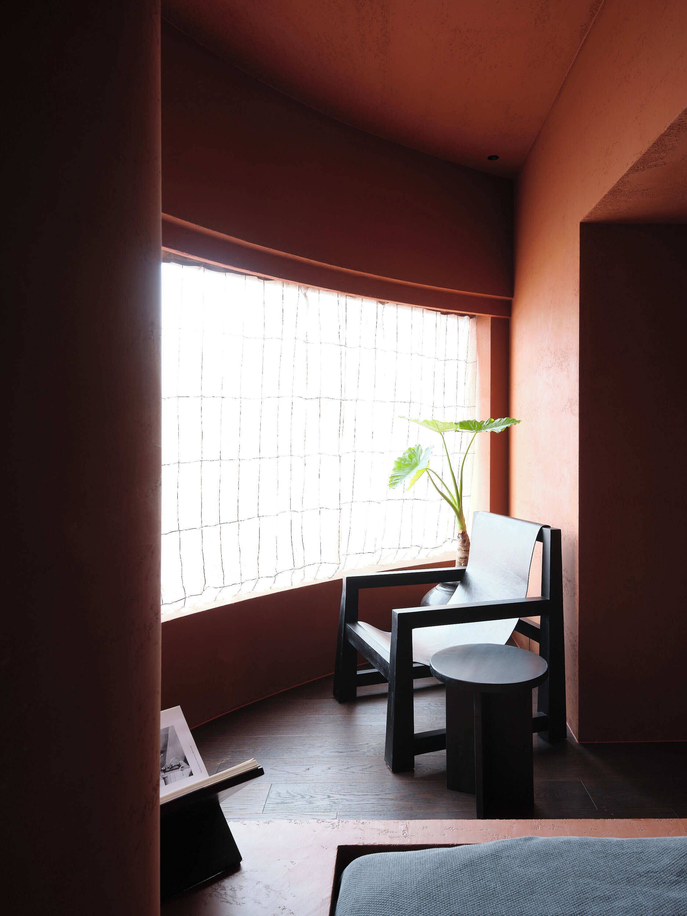 Um quarto pequeno e com pouca luz, com uma cadeira e uma mesinha. Uma janela com persianas permite a entrada de luz natural.