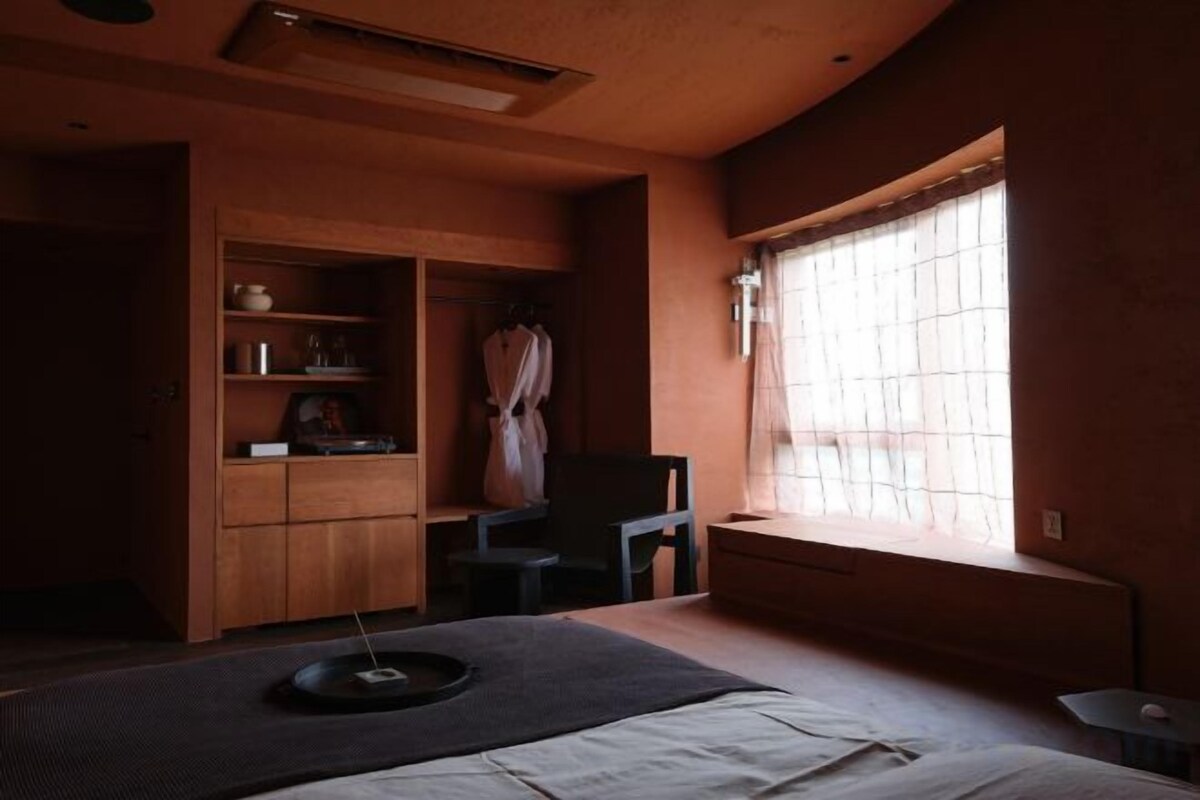 A room with a bed, a window with blinds, a chair, and a cabinet with shelves.