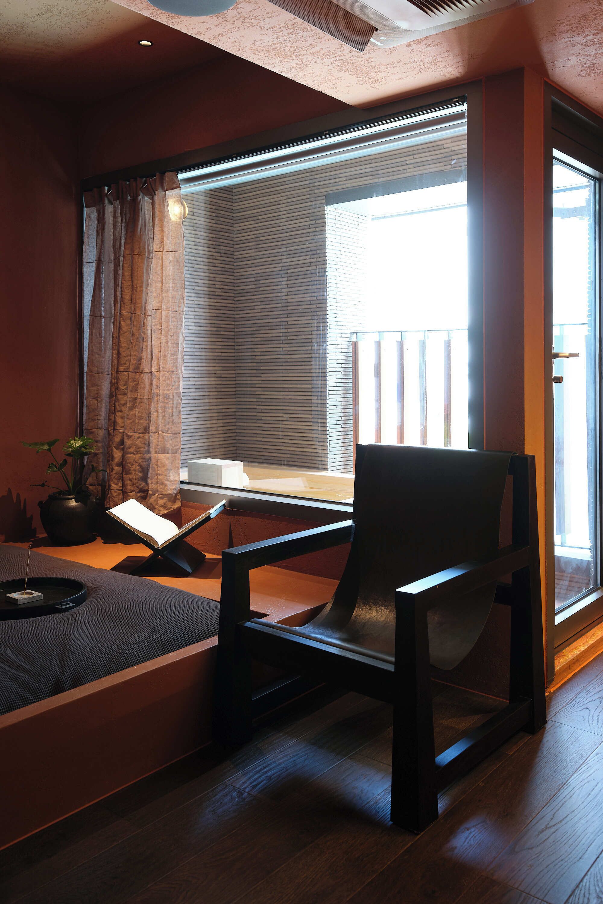 Una habitación con piso de madera, una silla negra, una ventana con persianas, una planta en maceta y una pequeña mesa con un libro.