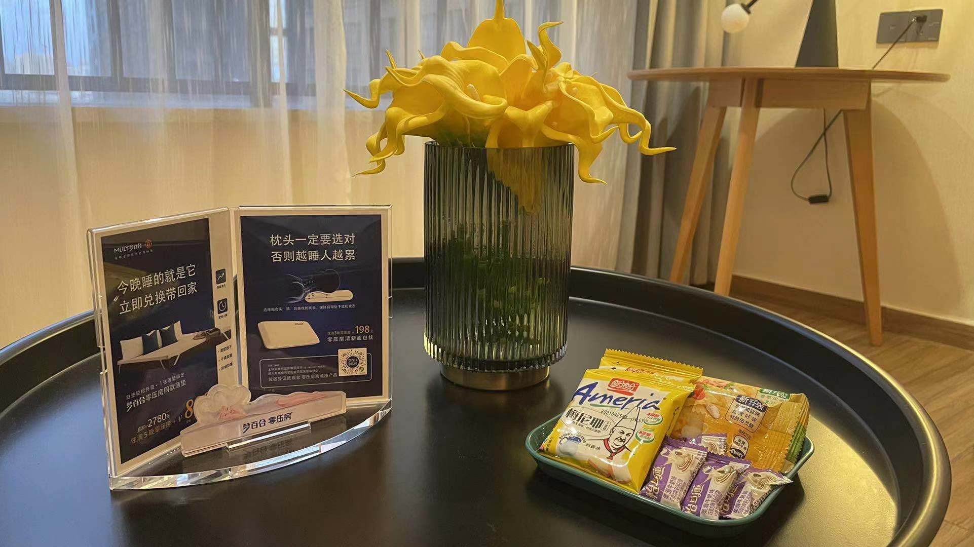 A hotel room with a brochure, a vase with yellow flowers, and a tray with packaged snacks.