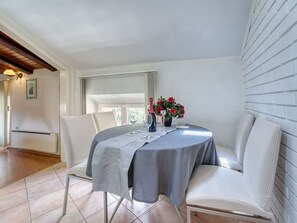 A dining area with a round table, white chairs, and a grey tablecloth. There are two red flowers in a vase on the table.