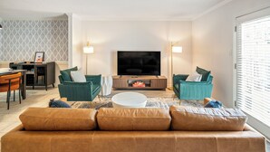 A living room with a brown sofa, teal armchairs, a TV, and a dining area with a table and chairs.