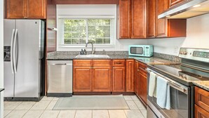 A kitchen with wooden cabinets, stainless steel appliances, a window with blinds, and a tiled floor.