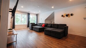 Un salon moderne avec un canapé d’angle, un téléviseur à écran plat fixé au mur et un plancher en bois.