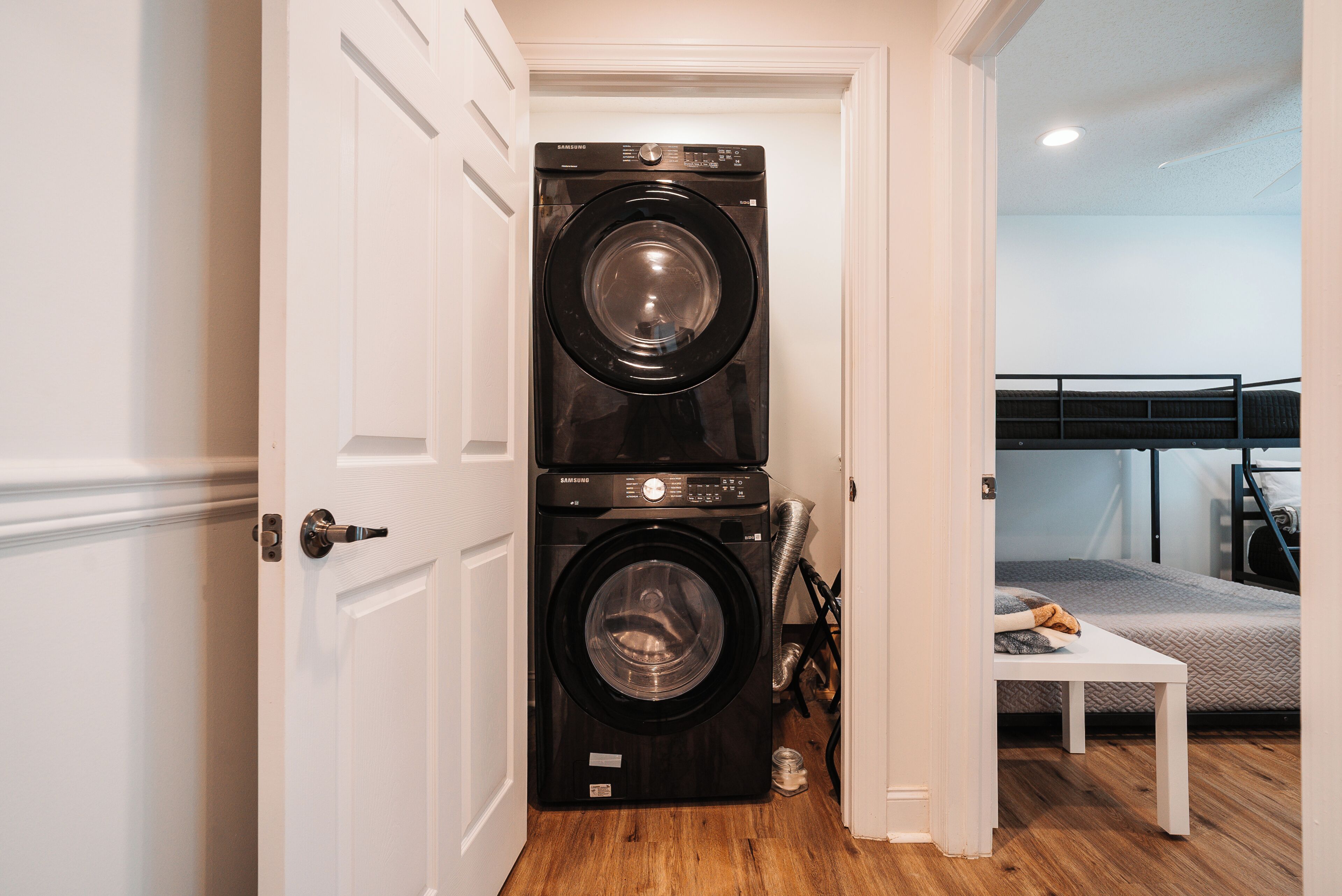 A laundry room with stacked washers and dryers, a bed, and a table in a bedroom.