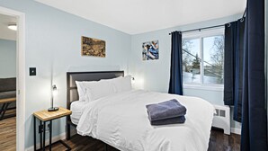 A bedroom with a bed, a bedside table, a window with curtains, and artwork on the walls. - 5 Min walk to restaurants/cafes, 8 min drive to airport, 10 min walk to trails. (Anchorage)